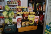 A display of various packaged food products is arranged on a wooden platform. The products include snack packs and containers in vibrant packaging with different colors such as green, yellow, and red. A decorative item resembling a small tree with gold and green leaves is placed alongside. Additionally, there is a small, dark green retro-style radio on the left side of the arrangement.