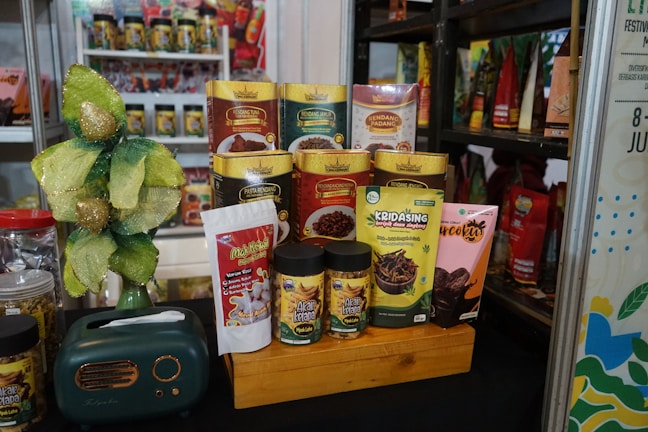 A display of various packaged food products is arranged on a wooden platform. The products include snack packs and containers in vibrant packaging with different colors such as green, yellow, and red. A decorative item resembling a small tree with gold and green leaves is placed alongside. Additionally, there is a small, dark green retro-style radio on the left side of the arrangement.