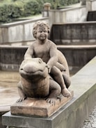 A stone statue depicting a cherubic child sitting atop a large frog, with the word 'SEX' inscribed on the forehead of the child figure. The sculpture is placed on a concrete base in what appears to be a park or garden setting.