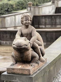 A stone statue depicting a cherubic child sitting atop a large frog, with the word 'SEX' inscribed on the forehead of the child figure. The sculpture is placed on a concrete base in what appears to be a park or garden setting.