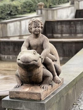 A stone statue depicting a cherubic child sitting atop a large frog, with the word 'SEX' inscribed on the forehead of the child figure. The sculpture is placed on a concrete base in what appears to be a park or garden setting.