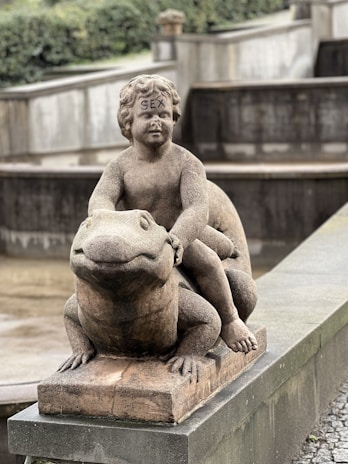 A stone statue depicting a cherubic child sitting atop a large frog, with the word 'SEX' inscribed on the forehead of the child figure. The sculpture is placed on a concrete base in what appears to be a park or garden setting.