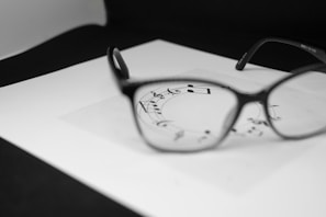A pair of eyeglasses is placed on a piece of white paper featuring musical notes. The glasses are in sharp focus, highlighting the notes seen through the lenses, while the rest of the image is gently blurred.