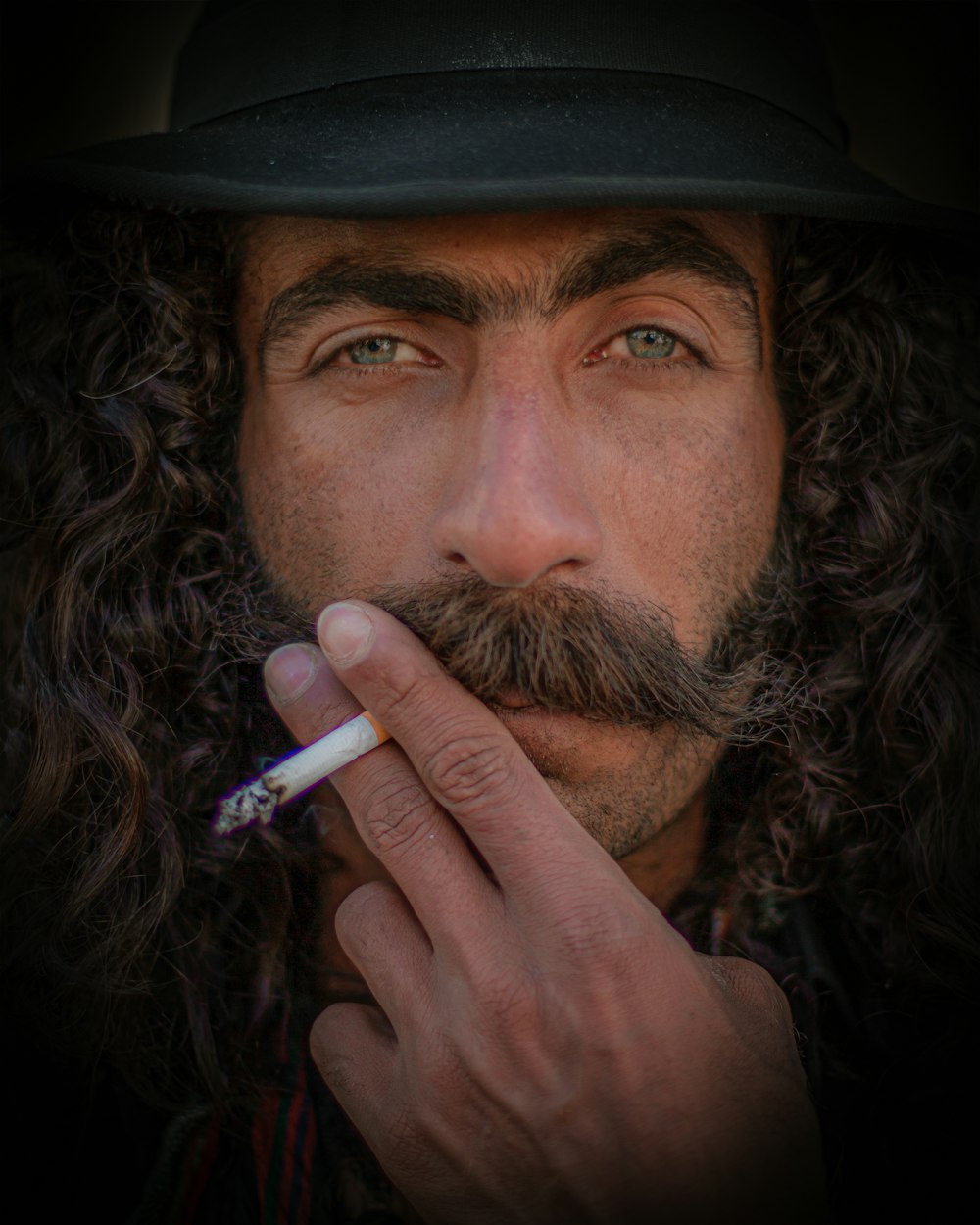 a man with long curly hair wearing a hat and holding a cigarette