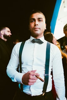 A man wearing a white dress shirt with blue suspenders and a bow tie holds a cigar. He has a serious expression and is looking slightly upward. The background includes other people, some of whom are partially visible. The lighting has a warm tone, giving the scene an elegant atmosphere.