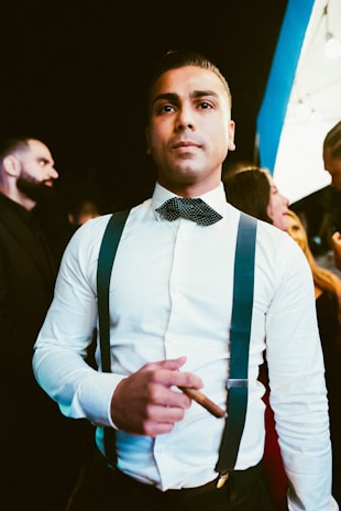 A man wearing a white dress shirt with blue suspenders and a bow tie holds a cigar. He has a serious expression and is looking slightly upward. The background includes other people, some of whom are partially visible. The lighting has a warm tone, giving the scene an elegant atmosphere.
