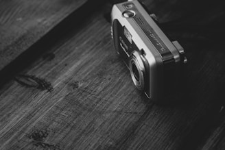 A classic digital camera is placed on a wooden surface. The image is in black and white, highlighting the textures of the camera and the wooden table.