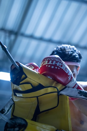 A boxer wearing red boxing gloves is punching a yellow punching bag with focus and intensity. The gloves have branding text on them, and the scene is set in a gym with a metallic roof structure in the background. The lighting creates a dramatic and energetic atmosphere.