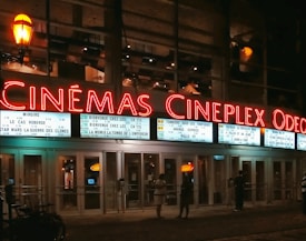 A night scene of a movie theater with bright neon signs displaying 'CINÉMAS CINEPLEX ODEON'. Movie listings are visible on the marquee along with showtimes. A few people are standing in front of the entrance, which is illuminated by the interior lights.