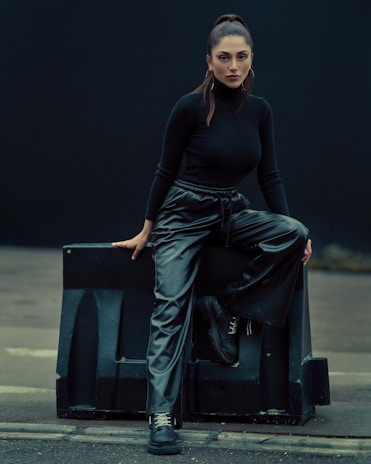 A person dressed in a stylish black ensemble consisting of a long-sleeved top and shiny black pants sits confidently on a large dark block. The setting has a subdued, almost industrial feel with muted lighting. The person's hair is neatly tied back, and they are wearing large hoop earrings.