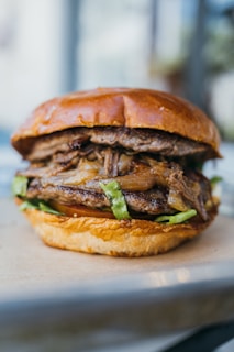 A juicy burger with multiple layers, including a glossy, golden brown bun on top. It features well-cooked meat, grilled onions, and lettuce, with a visible tomato and potentially cheese in the lower section. The focus is sharp, highlighting the textures and colors, with a blurred background suggesting a relaxed, possibly outdoor setting.
