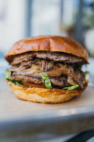 A juicy burger with multiple layers, including a glossy, golden brown bun on top. It features well-cooked meat, grilled onions, and lettuce, with a visible tomato and potentially cheese in the lower section. The focus is sharp, highlighting the textures and colors, with a blurred background suggesting a relaxed, possibly outdoor setting.