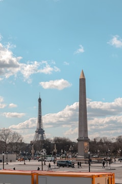 The image features a cityscape with an obelisk in the foreground and the Eiffel Tower in the background under a partly cloudy sky. People are walking around a public square, and a few cars are visible on the street.