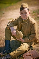 A young boy dressed in vintage military attire sits on the ground holding a canteen. He wears a brown uniform with matching cap and black boots. A leather notebook lies beside him on the grass, and the background is an outdoor setting.