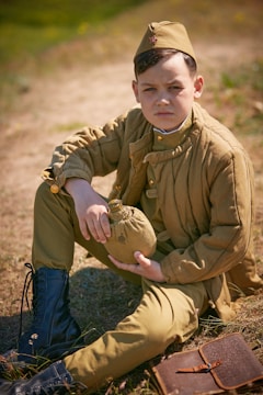 A young boy dressed in vintage military attire sits on the ground holding a canteen. He wears a brown uniform with matching cap and black boots. A leather notebook lies beside him on the grass, and the background is an outdoor setting.
