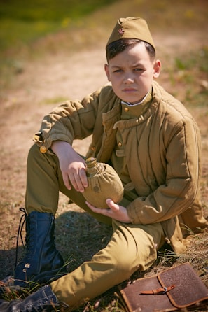A young boy dressed in vintage military attire sits on the ground holding a canteen. He wears a brown uniform with matching cap and black boots. A leather notebook lies beside him on the grass, and the background is an outdoor setting.