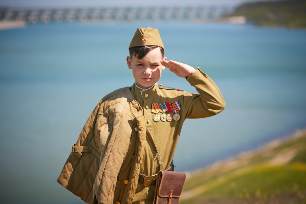 a boy in a uniform saluting in front of a body of water