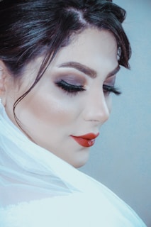 A close-up portrait of a person with makeup featuring bold red lipstick, dark eyeshadow, and long lashes, hair styled elegantly, and partially draped in a translucent white fabric.