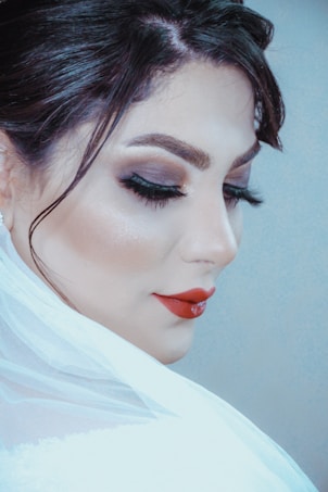 A close-up portrait of a person with makeup featuring bold red lipstick, dark eyeshadow, and long lashes, hair styled elegantly, and partially draped in a translucent white fabric.