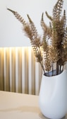 A sleek, white vase filled with dried plants is placed on a light-colored surface. The background features vertical stripes with soft lighting that creates an elegant and minimalist aesthetic.