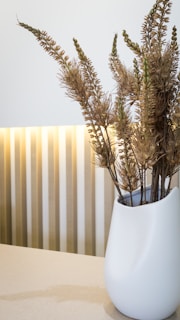 A sleek, modern white vase with clean lines sitting on a wooden shelf against a soft gray wall.