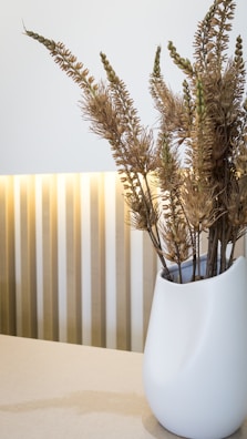 A sleek, white vase filled with dried plants is placed on a light-colored surface. The background features vertical stripes with soft lighting that creates an elegant and minimalist aesthetic.