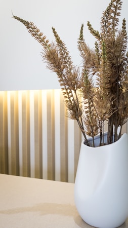 A sleek, white vase filled with dried plants is placed on a light-colored surface. The background features vertical stripes with soft lighting that creates an elegant and minimalist aesthetic.