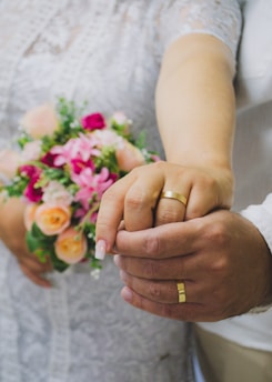 Two hands with wedding rings are joined together, one belongs to a bride in a lace dress holding a bouquet of pink and peach flowers.