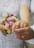 Two hands with wedding rings are joined together, one belongs to a bride in a lace dress holding a bouquet of pink and peach flowers.