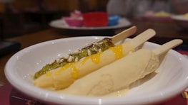 A white bowl contains ice cream or frozen desserts on sticks, one covered with green sauce and nut topping, and the other with a pale yellow sauce. In the background, a plate holds more unspecified items, adding a colorful and playful contrast.
