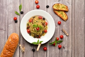 A wooden table setting features a white plate containing a dish of spaghetti garnished with cherry tomatoes, olives, and herbs. Surrounding the plate are additional cherry tomatoes, black olives, fresh basil leaves, two slices of garlic bread with parsley, and a loaf of bread. The rustic backdrop and scattered ingredients create a warm and inviting atmosphere.