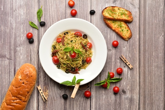 A wooden table setting features a white plate containing a dish of spaghetti garnished with cherry tomatoes, olives, and herbs. Surrounding the plate are additional cherry tomatoes, black olives, fresh basil leaves, two slices of garlic bread with parsley, and a loaf of bread. The rustic backdrop and scattered ingredients create a warm and inviting atmosphere.