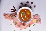 A white bowl filled with a rich, creamy orange-brown curry, garnished with cream and a sprig of cilantro, surrounded by spices. There are star anise, red chilies, onion rings, a lemon wedge, and fresh mint leaves placed artfully on a light textured surface. The bowl is placed atop a soft, pinkish-orange cloth.