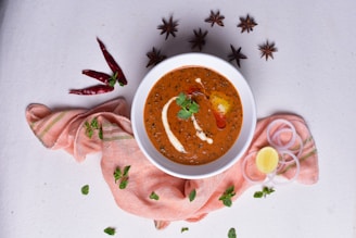 A white bowl filled with a rich, creamy orange-brown curry, garnished with cream and a sprig of cilantro, surrounded by spices. There are star anise, red chilies, onion rings, a lemon wedge, and fresh mint leaves placed artfully on a light textured surface. The bowl is placed atop a soft, pinkish-orange cloth.