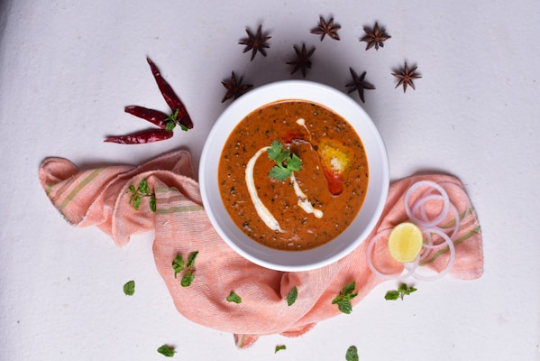 A white bowl filled with a rich, creamy orange-brown curry, garnished with cream and a sprig of cilantro, surrounded by spices. There are star anise, red chilies, onion rings, a lemon wedge, and fresh mint leaves placed artfully on a light textured surface. The bowl is placed atop a soft, pinkish-orange cloth.