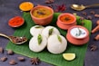 A traditional South Indian meal is presented on a banana leaf, featuring fluffy white idlis garnished with seasoning. Accompanying the idlis are three types of chutneys and sambar served in small clay pots. The setting includes a variety of spices, such as star anise and black cardamom, along with red chili powder and turmeric displayed in small bowls. A wedge of lime is placed on the leaf, enhancing the colorful and aromatic presentation.