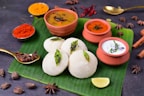 A traditional South Indian meal is presented on a banana leaf, featuring fluffy white idlis garnished with seasoning. Accompanying the idlis are three types of chutneys and sambar served in small clay pots. The setting includes a variety of spices, such as star anise and black cardamom, along with red chili powder and turmeric displayed in small bowls. A wedge of lime is placed on the leaf, enhancing the colorful and aromatic presentation.