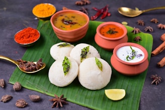 A traditional South Indian meal is presented on a banana leaf, featuring fluffy white idlis garnished with seasoning. Accompanying the idlis are three types of chutneys and sambar served in small clay pots. The setting includes a variety of spices, such as star anise and black cardamom, along with red chili powder and turmeric displayed in small bowls. A wedge of lime is placed on the leaf, enhancing the colorful and aromatic presentation.