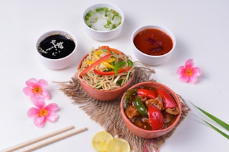 An arrangement of a meal featuring colorful stir-fried noodles topped with sliced red and yellow bell peppers, served in a bowl, alongside a bowl of saucy stir-fried vegetables with red and green peppers. Accompanying the main dishes are three dipping sauces in small white bowls: a black sesame sauce, a clear soup with green onions, and a red chili sauce. The setting includes decorative pink flowers, lemon slices, and chopsticks on a white background, creating a vibrant and appetizing presentation.