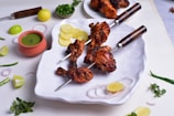 A plate with grilled chicken skewers placed on a white dish, decorated with slices of lemon and onion. A small pot of green chutney is nearby, along with lemon pieces, mint leaves, and green chilies scattered around. The chicken is charred and spiced, giving it an appealing look.