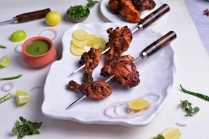 A plate with grilled chicken skewers placed on a white dish, decorated with slices of lemon and onion. A small pot of green chutney is nearby, along with lemon pieces, mint leaves, and green chilies scattered around. The chicken is charred and spiced, giving it an appealing look.