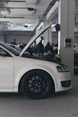 A white car with its hood open parked inside a large garage. Several people are gathered in the background, indicating a possible gathering or event. The environment appears industrial with exposed beams and fluorescent lighting.
