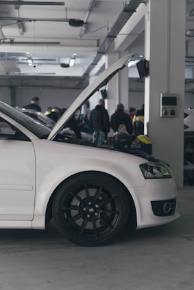 A white car with its hood open parked inside a large garage. Several people are gathered in the background, indicating a possible gathering or event. The environment appears industrial with exposed beams and fluorescent lighting.
