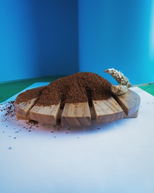 A wooden platform holds a mound of brown granular material, likely coffee grounds. A decorative wheat stem is placed on the right side. The background is a gradient of blue and green, providing a clean and vibrant contrast.