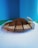 A wooden platform holds a mound of brown granular material, likely coffee grounds. A decorative wheat stem is placed on the right side. The background is a gradient of blue and green, providing a clean and vibrant contrast.