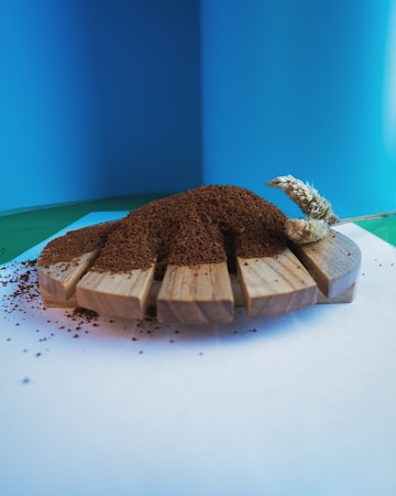 A wooden platform holds a mound of brown granular material, likely coffee grounds. A decorative wheat stem is placed on the right side. The background is a gradient of blue and green, providing a clean and vibrant contrast.