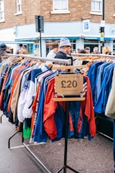A street market scene featuring a clothing rack filled with colorful jackets, marked with a price tag of £20, and labeled 'Broadway Vintage'. Several people are browsing the items on display. The background includes a brick building and a shop with a blue facade.