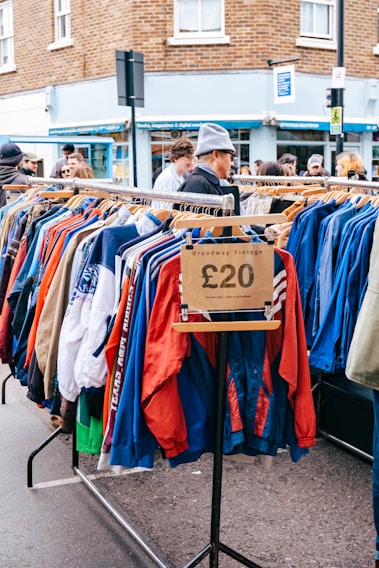A street market scene featuring a clothing rack filled with colorful jackets, marked with a price tag of £20, and labeled 'Broadway Vintage'. Several people are browsing the items on display. The background includes a brick building and a shop with a blue facade.