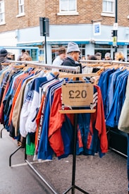 A street market scene featuring a clothing rack filled with colorful jackets, marked with a price tag of &pound;20, and labeled 'Broadway Vintage'. Several people are browsing the items on display. The background includes a brick building and a shop with a blue facade.