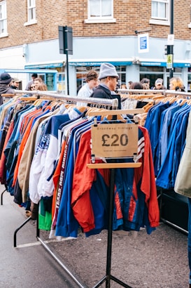 A street market scene featuring a clothing rack filled with colorful jackets, marked with a price tag of &pound;20, and labeled 'Broadway Vintage'. Several people are browsing the items on display. The background includes a brick building and a shop with a blue facade.